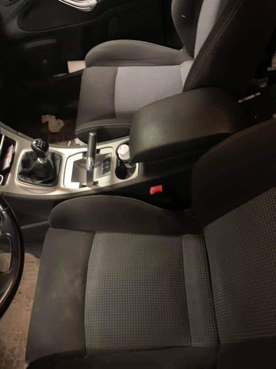 Wnętrze samochodu z widocznymi fotelami z szarą tapicerką, konsola środkowa z dźwignią zmiany biegów i podłokietnikiem, uchwyt na napoje.