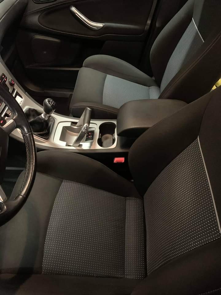 Wnętrze samochodu z ciemną tapicerką, widoczne fotele z szarymi wstawkami, dźwignia zmiany biegów i podłokietnik po zabiegu czyszczenia.