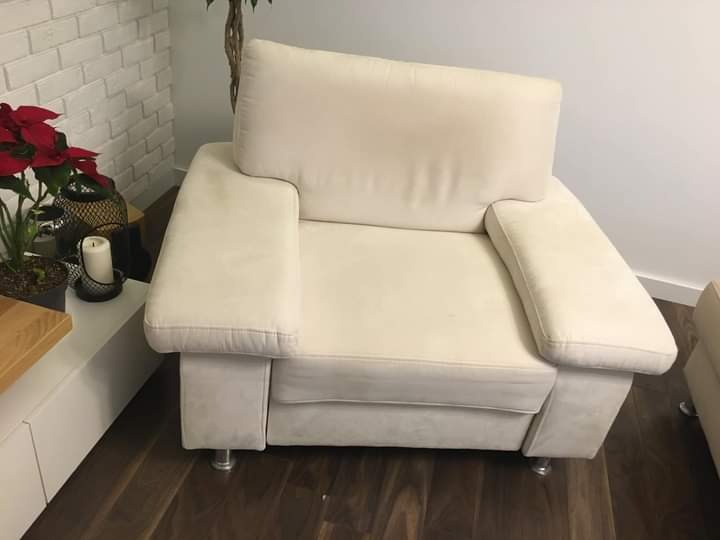 Jasny, beżowy fotel tapicerowany z podłokietnikami, ustawiony na ciemnej drewnianej podłodze, z widocznymi metalowymi nóżkami i rośliną poinsecji w tle.