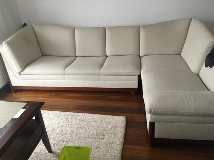 Jasny, narożny tapicerowany mebel wypoczynkowy z drewnianą podstawą, ustawiony na drewnianej podłodze obok stolika kawowego i kremowego dywanu.