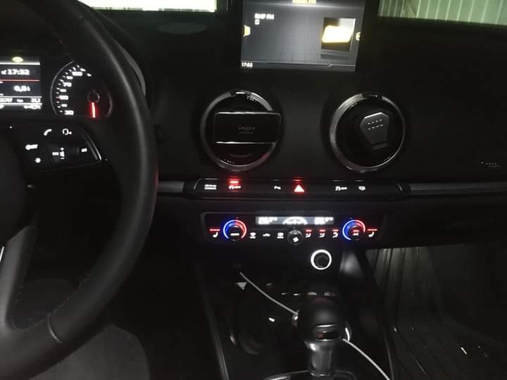 Wnętrze samochodu Audi A3, widoczny kokpit z włączonymi światłami i ekranem nawigacji, kierownica po lewej stronie, dźwignia zmiany biegów na pierwszym planie, ciemna kolorystyka.