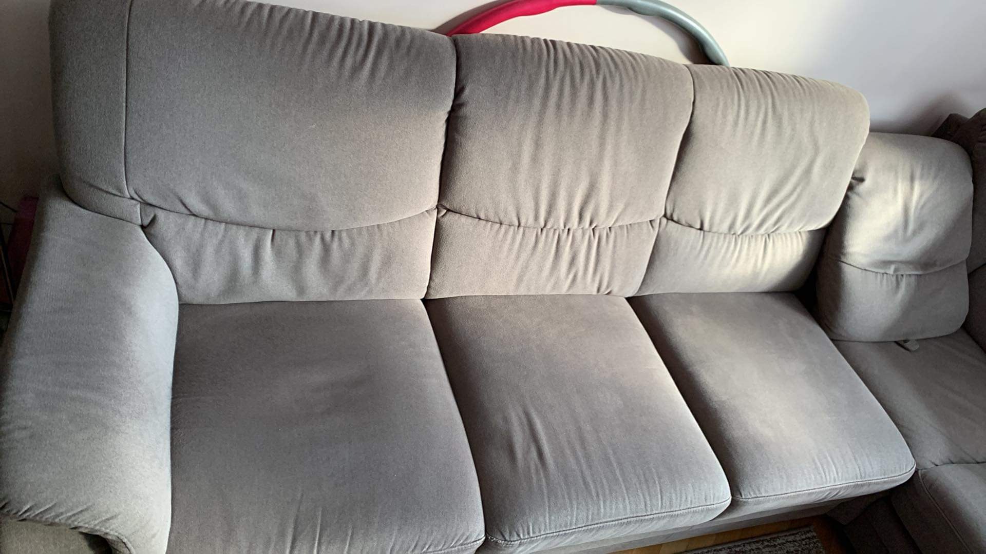 Szara, modułowa kanapa narożna z widocznymi śladami użytkowania i hula-hoop w tle, sugerująca domowe otoczenie.