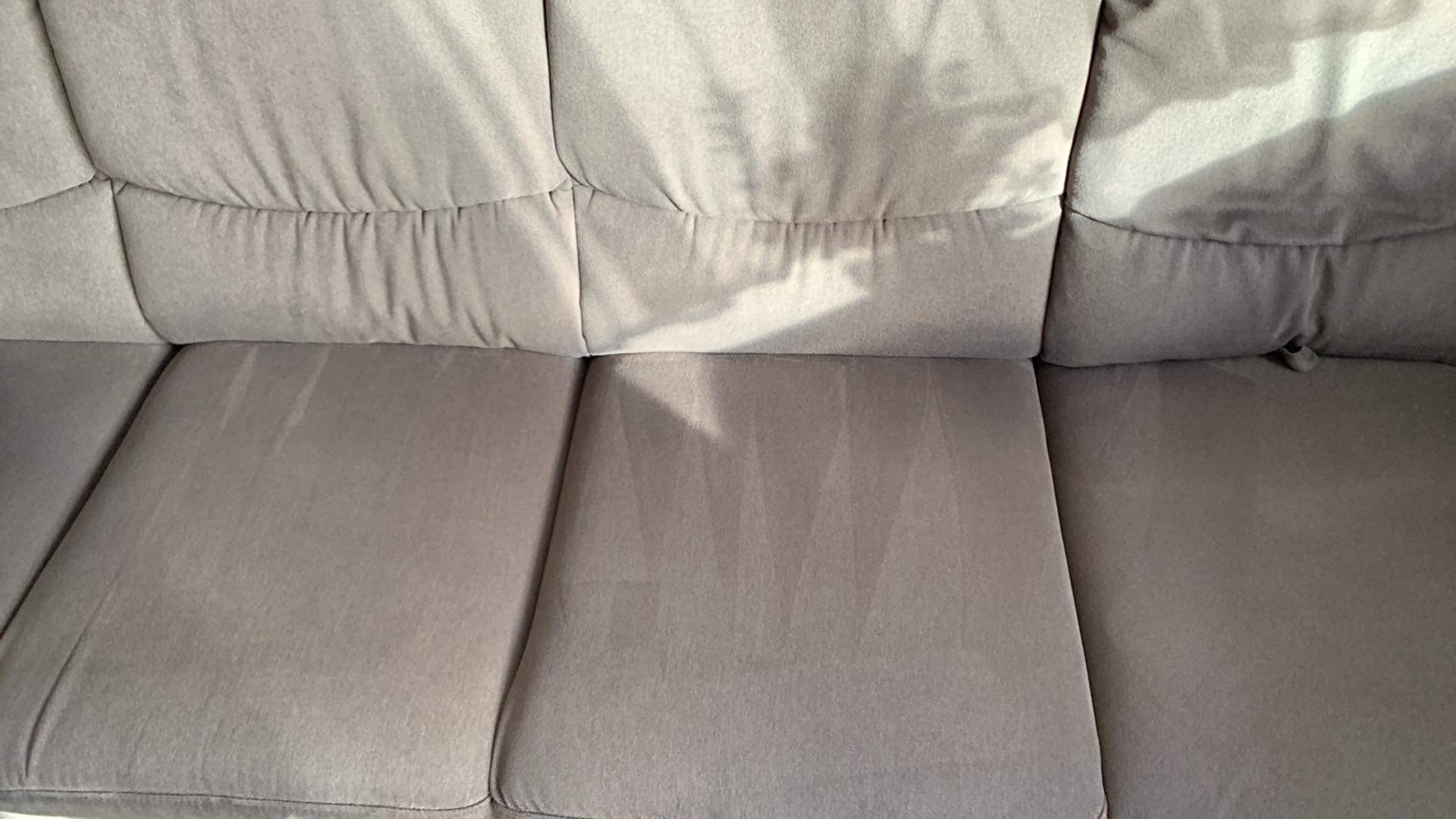 Szara sofa z widocznymi śladami po czyszczeniu, gdzie jaśniejsze fragmenty wskazują na obszary poddane niedawnemu praniu.