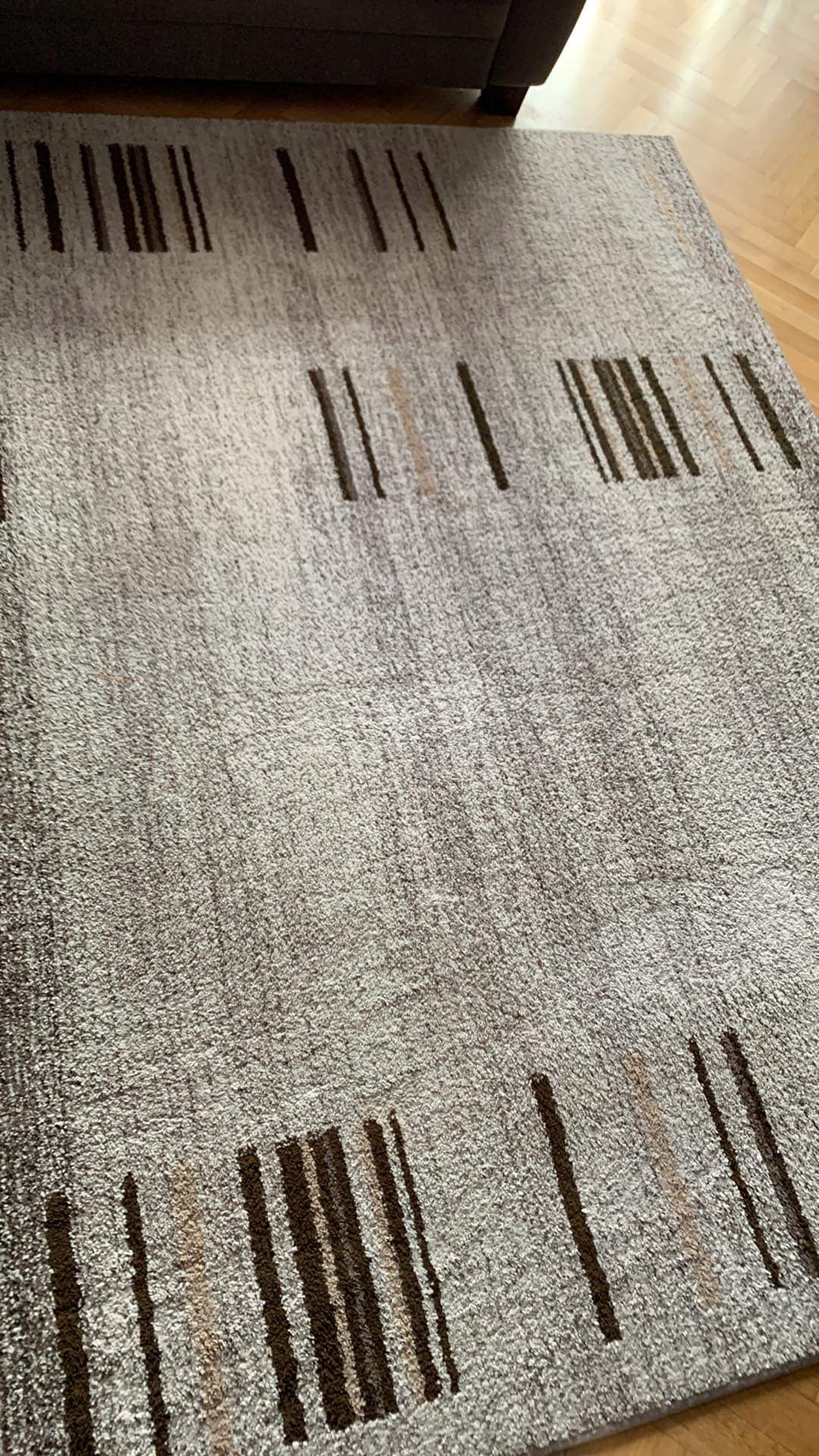 Szary dywan z abstrakcyjnym wzorem brązowych i beżowych pasków, leżący na drewnianej podłodze, fragment sofy widoczny w tle.