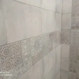 Jasne, matowe płytki ceramiczne w kabinie prysznicowej, z dekoracyjnym pasem płytek z motywami kwiatowymi i geometrycznymi, ułożone poziomo na ścianie.