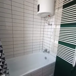 Biała wanna obudowana płytkami, grzejnik drabinkowy i bojler w łazience wykończonej w minimalistycznym stylu z płytkami imitującymi cegłę.