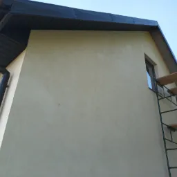 Ściana budynku z nałożonym tynkiem strukturalnym w odcieniach beżu, widoczne rusztowanie i orynnowanie w kolorze grafitowym.