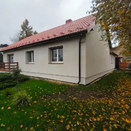 Ocieplony dom jednorodzinny z czerwoną dachówką i szarą elewacją, widoczny z boku na tle zielonego trawnika usłanego opadłymi liśćmi.