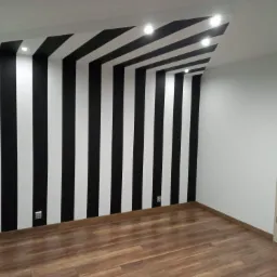 Pokój po remoncie z podłogą z paneli i ścianami w czarno-białe pionowe pasy, oświetlenie punktowe na suficie i jasne drzwi z przeszkleniami.