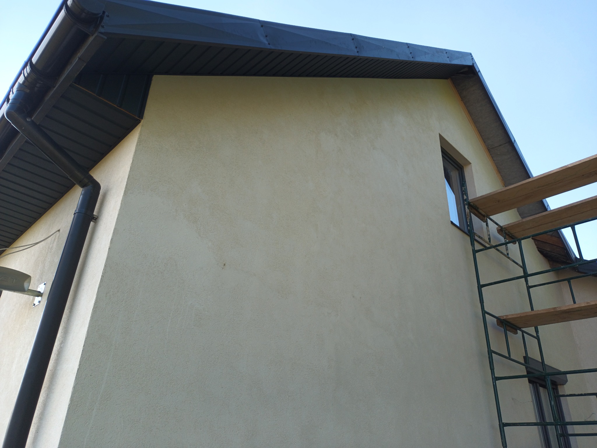 Ściana budynku z nałożonym tynkiem strukturalnym w odcieniach beżu, widoczne rusztowanie i orynnowanie w kolorze grafitowym.