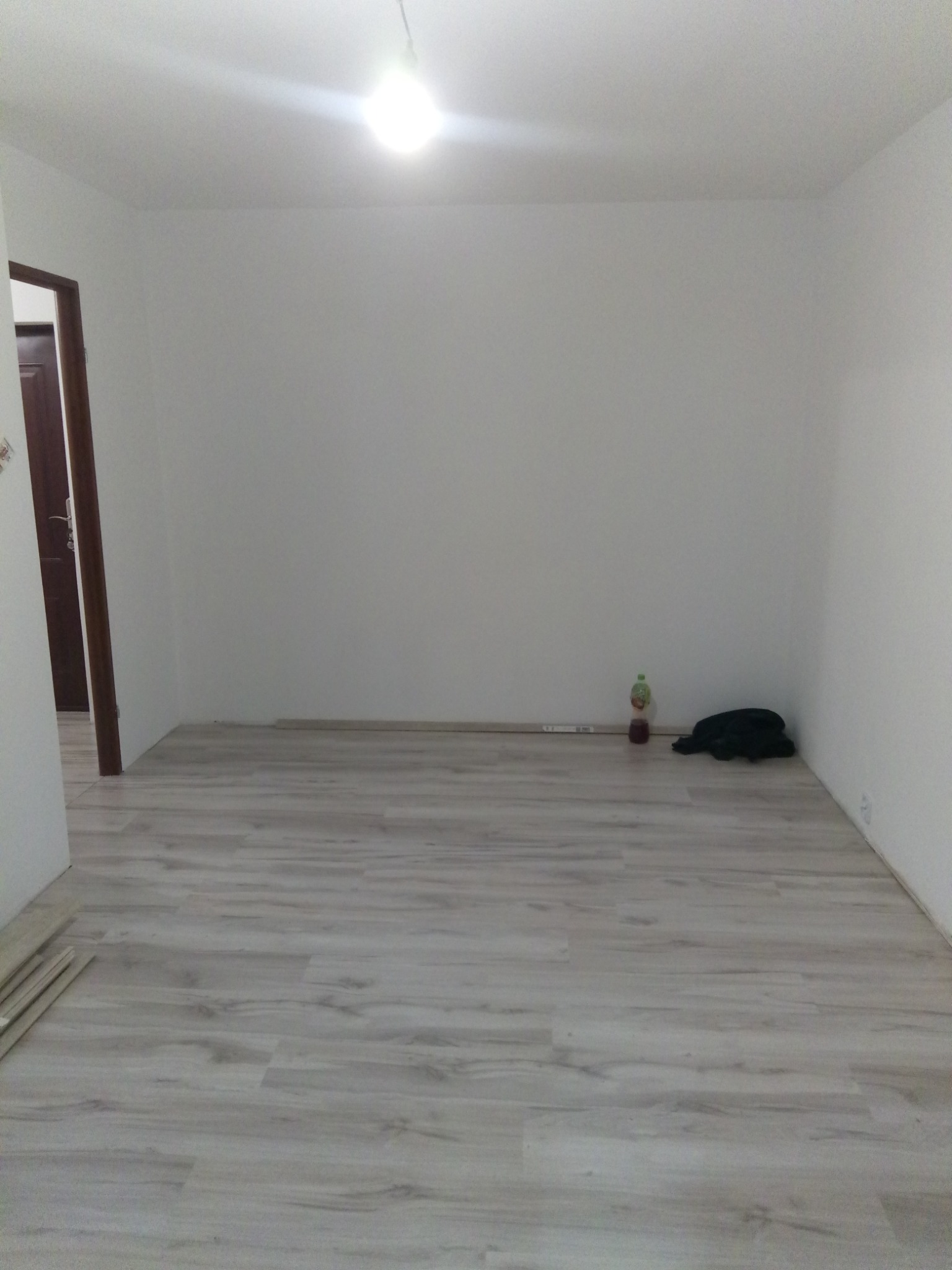 Pusty pokój po malowaniu ścian na biało i ułożeniu paneli podłogowych w odcieniach szarości. Widoczna butelka soku i czarna torba w rogu pokoju.