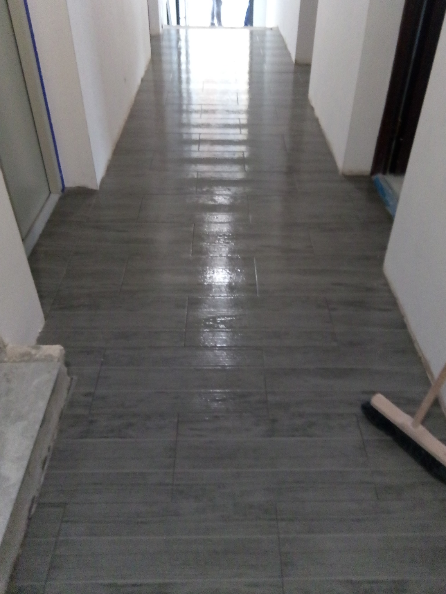 Świeżo ułożona, błyszcząca, szara podłoga z płytek imitujących drewno w długim korytarzu, z widoczną szczotką do zamiatania w rogu.