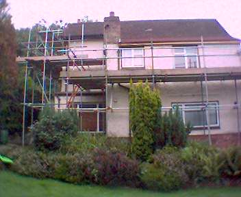 Dom jednorodzinny w trakcie remontu elewacji, otoczony rusztowaniem i zielenią. Widoczne krzewy i trawnik. Część dachu została już odnowiona.