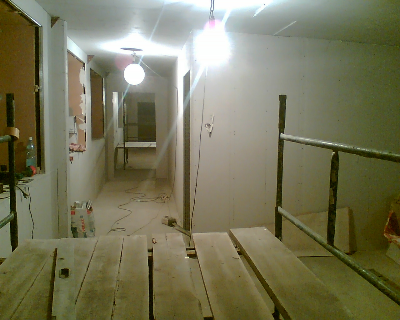 Wnętrze w trakcie remontu: widoczne rusztowania, płyty kartonowo-gipsowe na ścianach, sufit podwieszany w trakcie montażu, przewody elektryczne na podłodze, słabe oświetlenie.