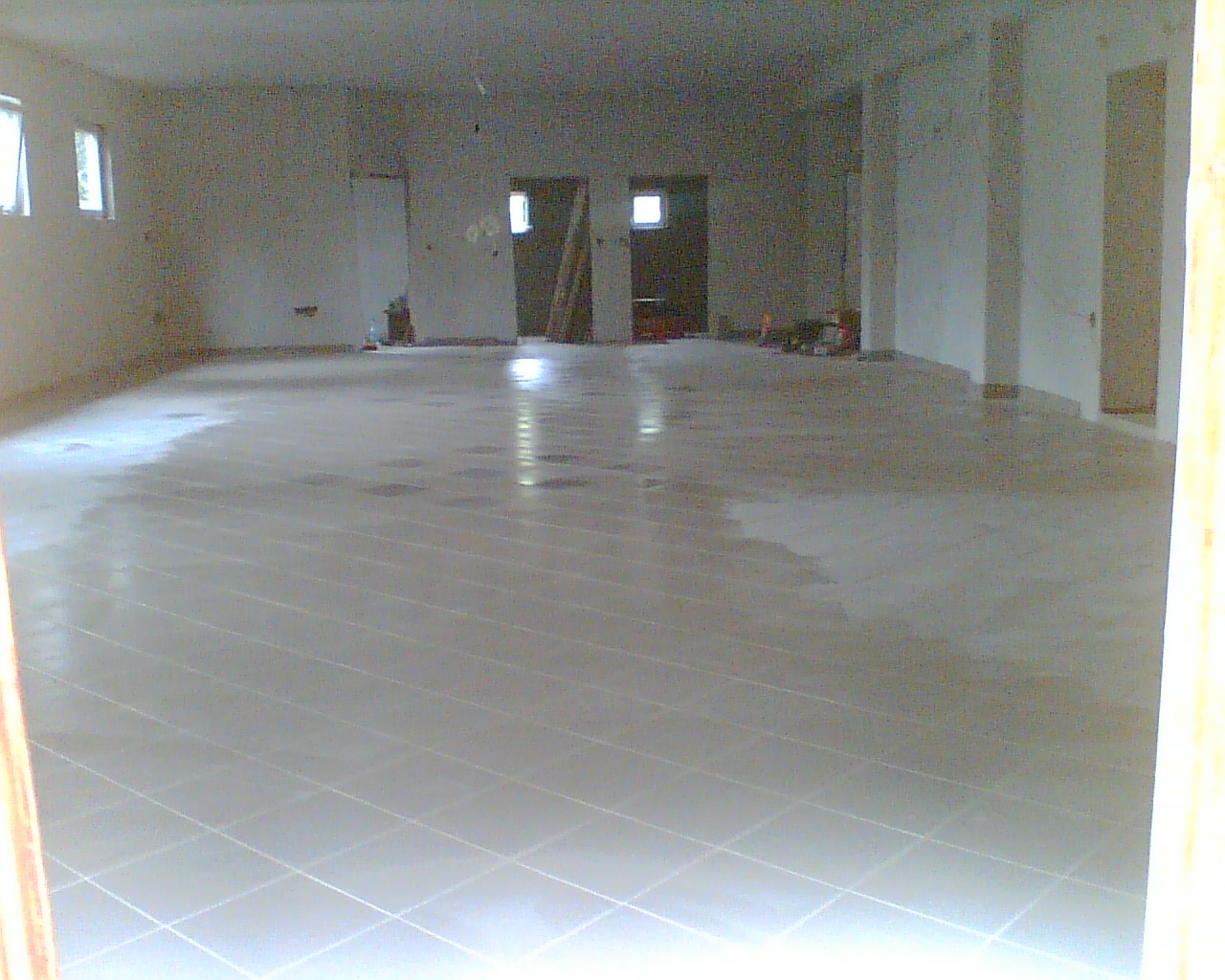 Wnętrze w trakcie wykańczania z nowo położonymi jasnymi płytkami podłogowymi, widoczne ślady wilgoci na powierzchni, surowe ściany i filary w tle, częściowo widoczne okna i drzwi.