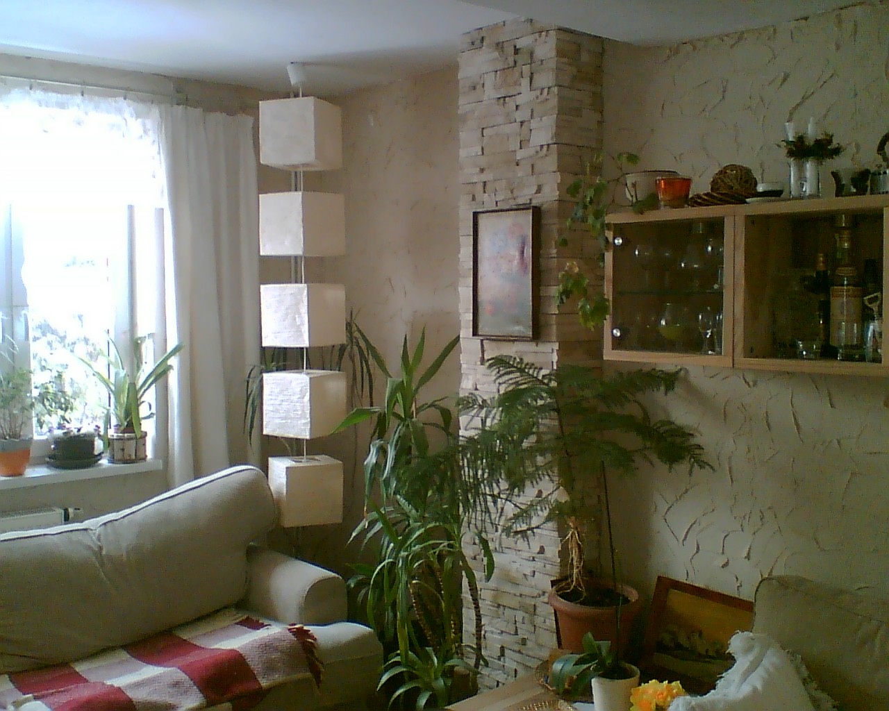 Jasne wnętrze pokoju z dekoracyjnym słupem z kamienia, wiszącą lampą z kwadratowych abażurów, roślinami doniczkowymi i teksturowaną ścianą