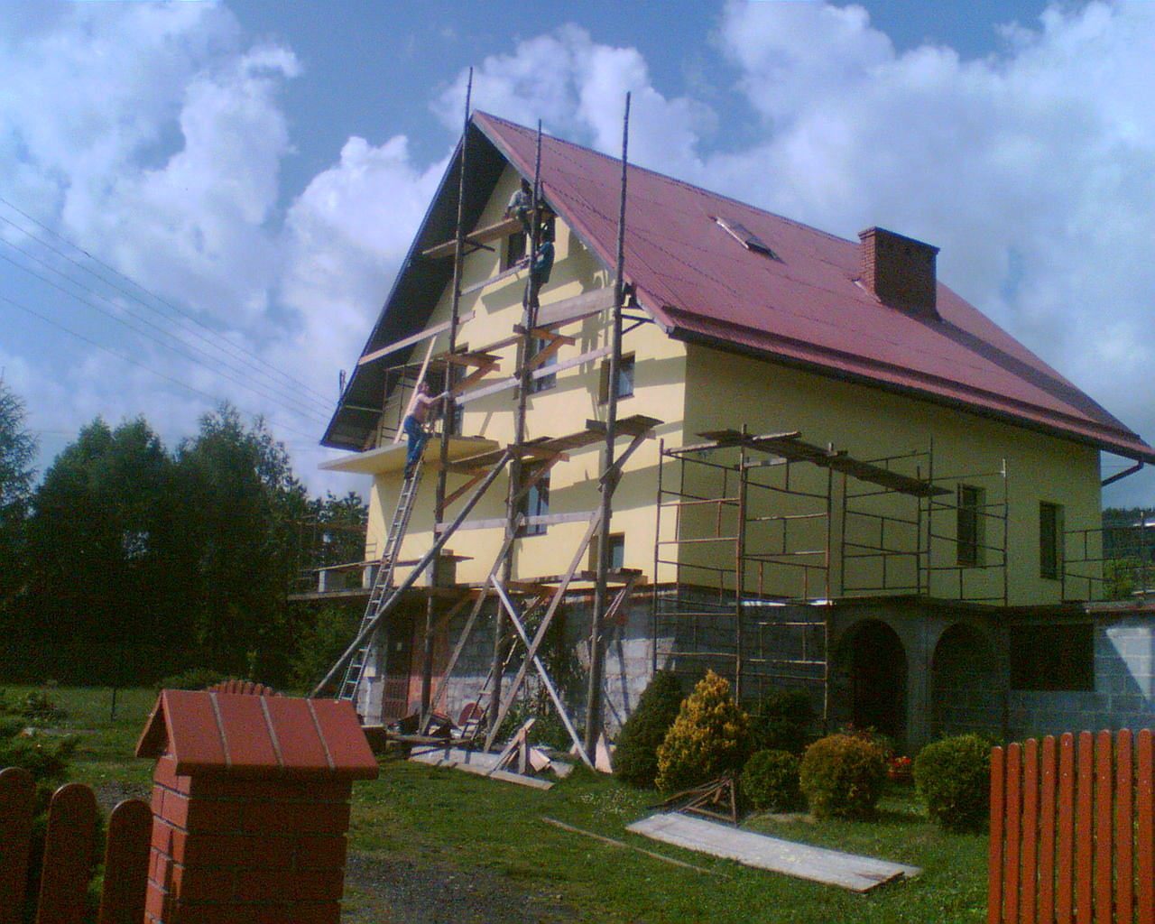 Dom w trakcie remontu elewacji, z widocznymi rusztowaniami, drabina oparta o ścianę, pracownicy na rusztowaniach, czerwony dach i częściowo wykończony fundament.