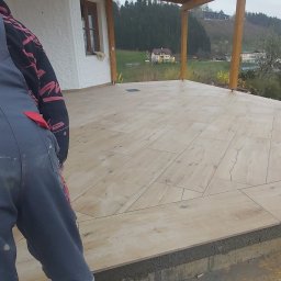 Kalmat - Układanie płytek imitujących drewno na tarasie zewnętrznym, widoczny glazurnik w roboczym ubraniu, w tle dom i pagórkowaty krajobraz.