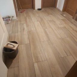 Kalmat - Ułożona jasna, drewnopodobna podłoga z płytek ceramicznych w korytarzu, widoczne trzy drewniane drzwi, wiadro z akcesoriami do układania płytek przy ścianie.