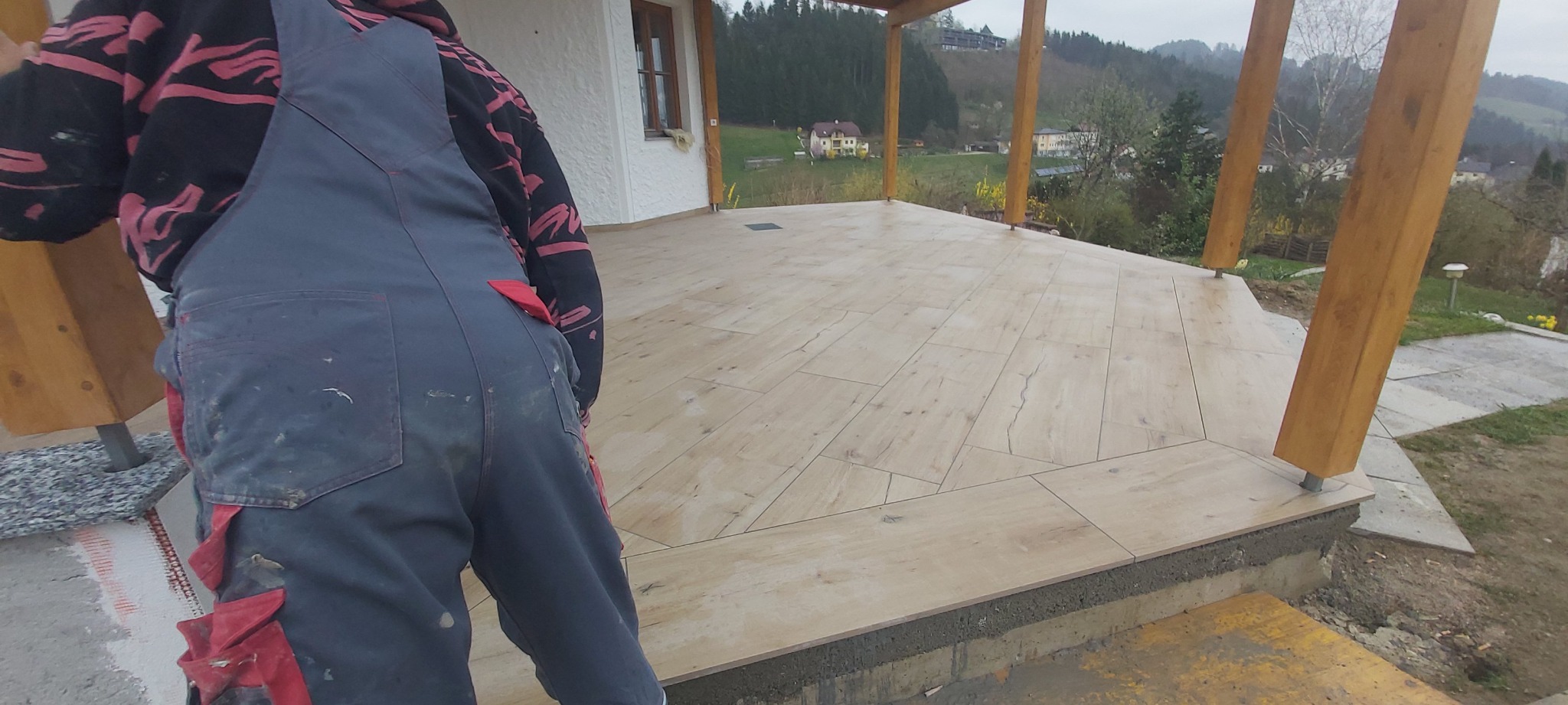 Układanie płytek imitujących drewno na tarasie zewnętrznym, widoczny glazurnik w roboczym ubraniu, w tle dom i pagórkowaty krajobraz.