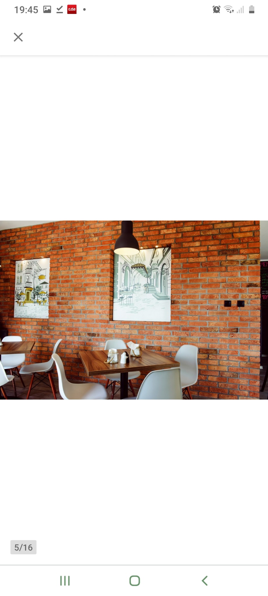 Ceglana ściana z dwoma obrazami przedstawiającymi uliczne sceny kawiarniane, oświetlona wiszącą lampą, z nowoczesnymi białymi krzesłami i drewnianymi stolikami w kawiarni.