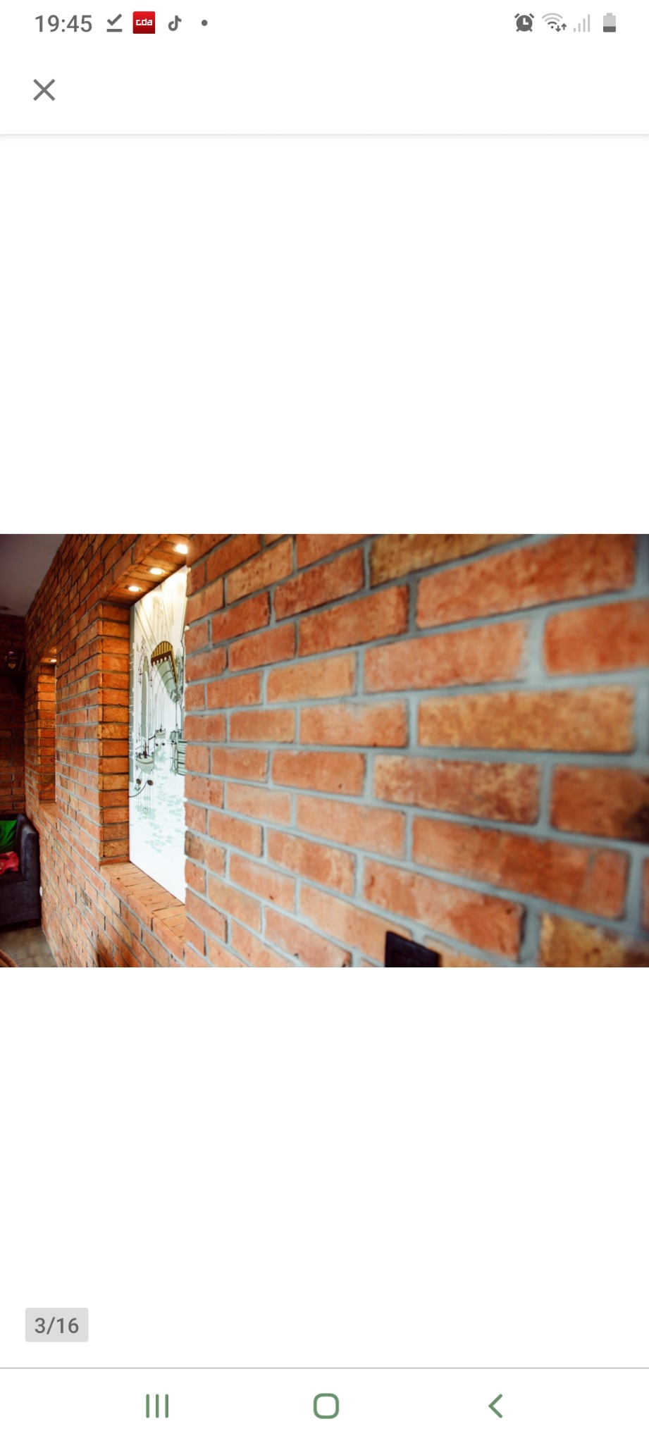 Ściana z czerwonej cegły klinkierowej wewnątrz pomieszczenia, widok z bliska, fragment ściany z oknem, w tle widoczne oświetlenie sufitowe i fragment wnętrza.