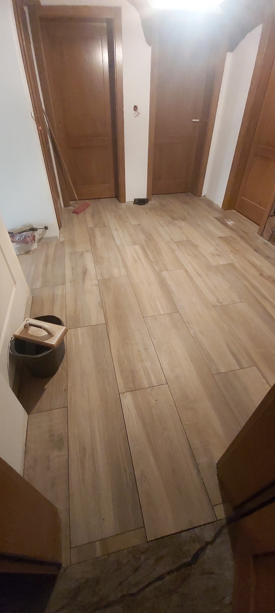 Ułożona jasna, drewnopodobna podłoga z płytek ceramicznych w korytarzu, widoczne trzy drewniane drzwi, wiadro z akcesoriami do układania płytek przy ścianie.