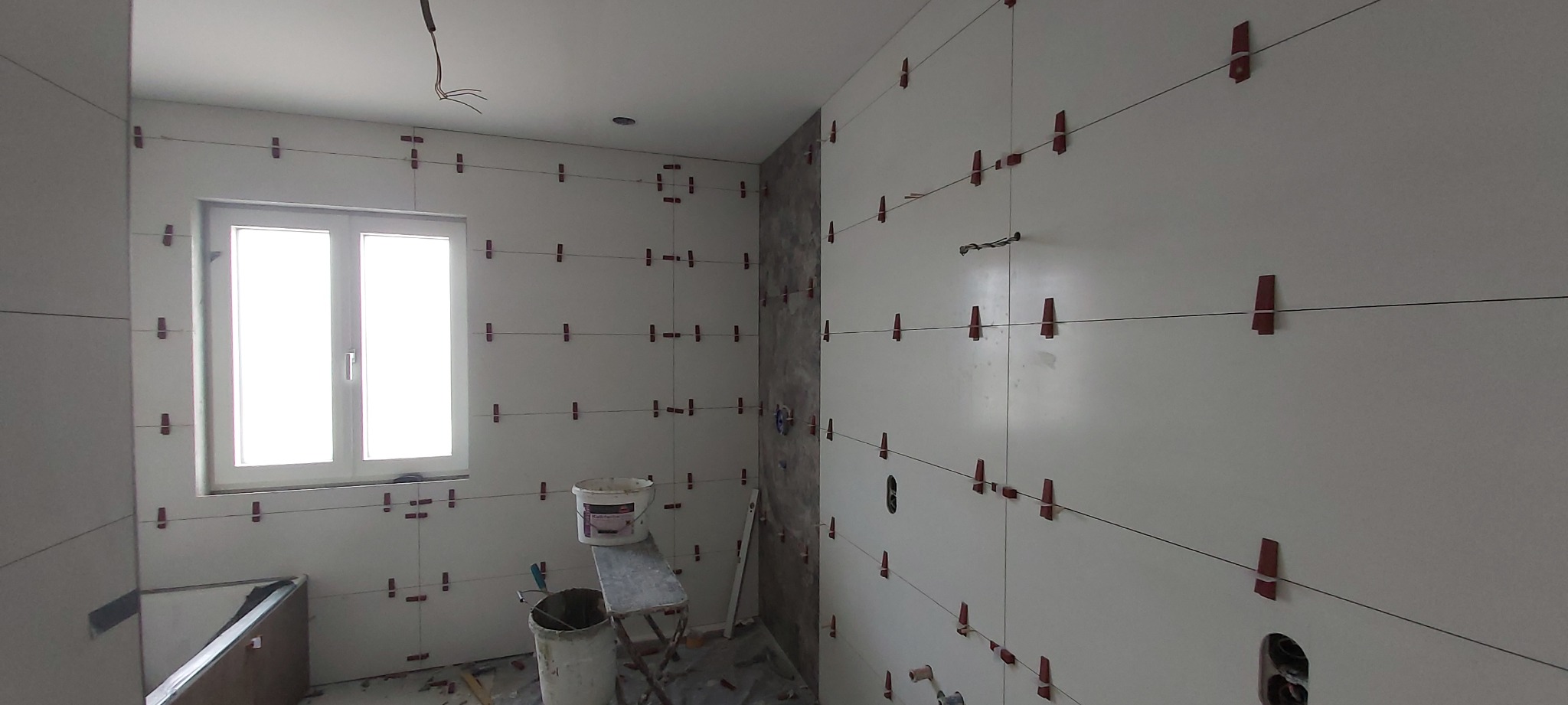 Białe płytki ścienne w trakcie układania, z czerwonymi krzyżykami dystansowymi i poziomymi liniami, widoczne wiaderka z zaprawą na tle okna.