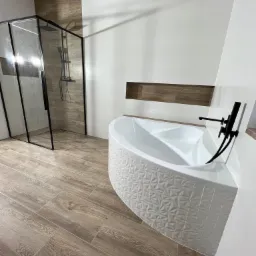 Nowoczesna łazienka z narożną wanną o trójwymiarowej fakturze, czarną armaturą i kabiną prysznicową typu walk-in z czarnymi profilami, wykończona płytkami imitującymi drewno i białymi płytkami.