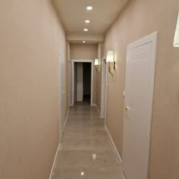 Korytarz w domu modułowym z beżowymi ścianami, marmurową podłogą i białymi drzwiami, oświetlony lampami ściennymi i sufitowymi.