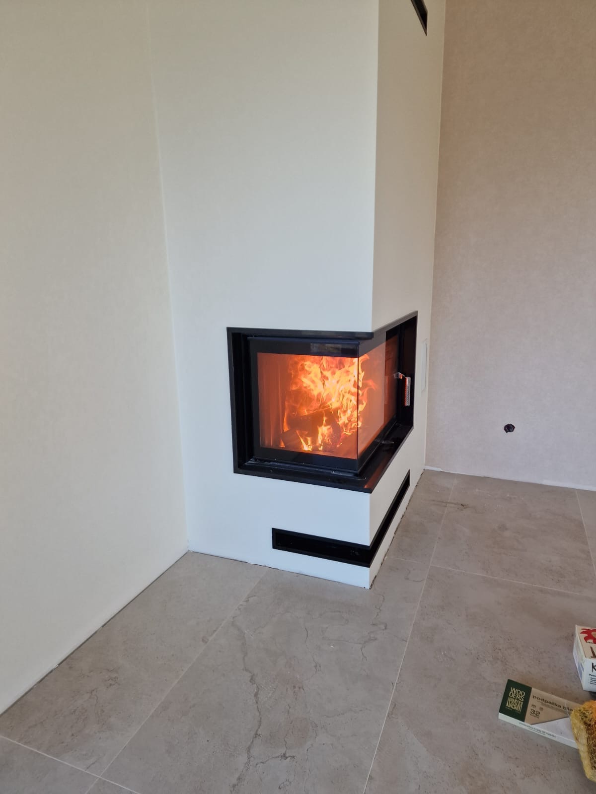 Nowoczesny narożny kominek z paleniskiem, obudowany białą konstrukcją z czarnymi akcentami, widoczny na tle jasnych ścian i podłogi z płytek imitujących marmur.