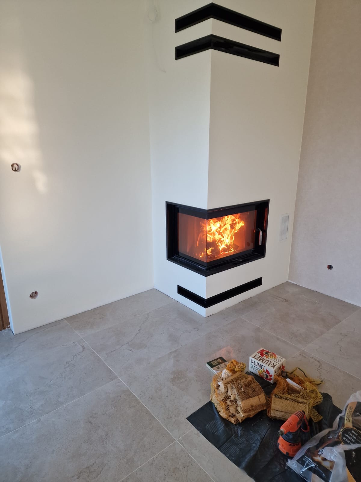 Nowoczesny narożny kominek z czarną ramą, otwarty ogień, białe ściany z czarnymi wstawkami, na podłodze jasne płytki, obok kominka drewno do palenia, w tle widoczne niedokończone wykończenia ścian.