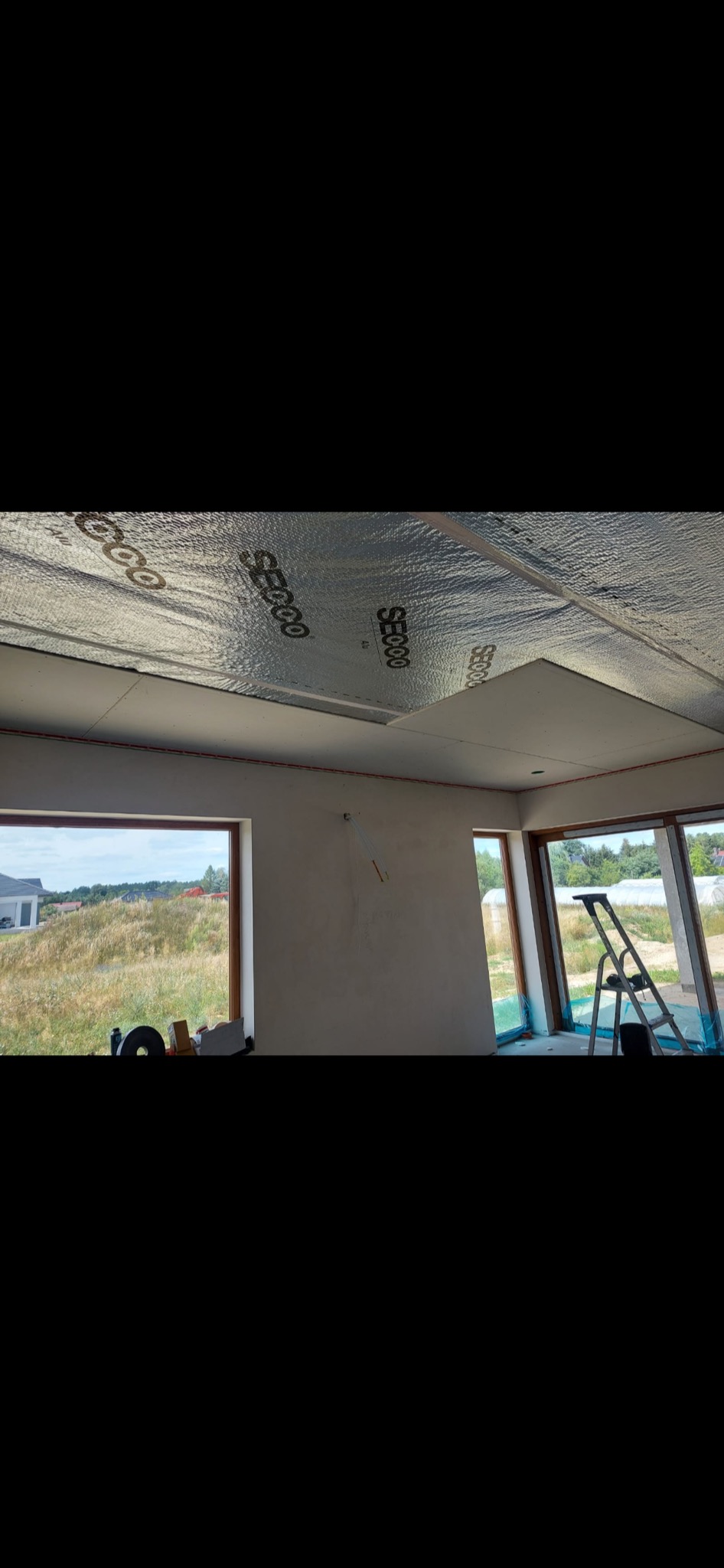 Wnętrze w trakcie budowy z widoczną izolacją termiczną SECOO na suficie, częściowo zamontowaną płytą gipsowo-kartonową, oknami z widokiem na zielony krajobraz i drabiną opartą o ścianę.