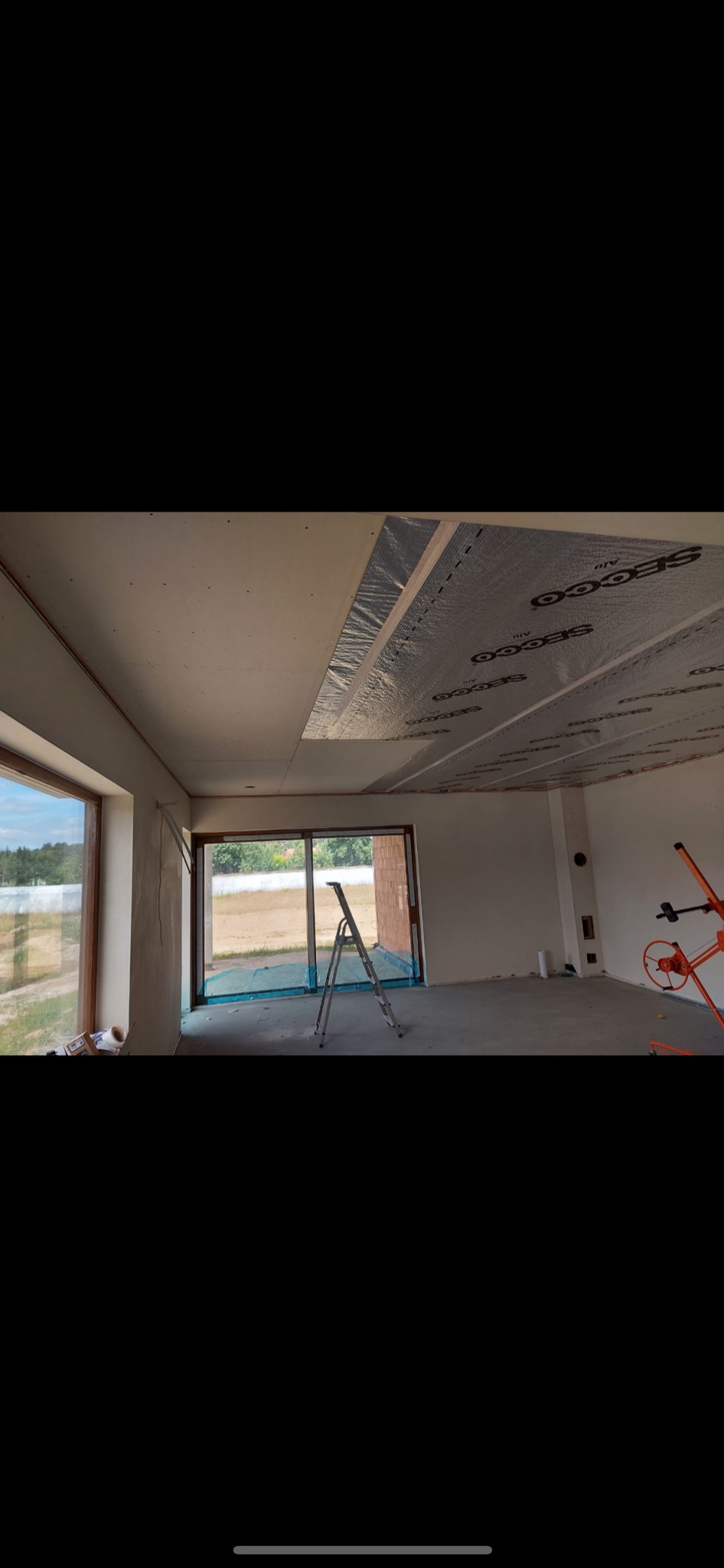 Wnętrze domu w trakcie wykończenia: widoczne płyty gipsowo-kartonowe na suficie z warstwą izolacji termicznej marki Secoo, drabina aluminiowa oraz mieszadło do zaprawy w narożniku.