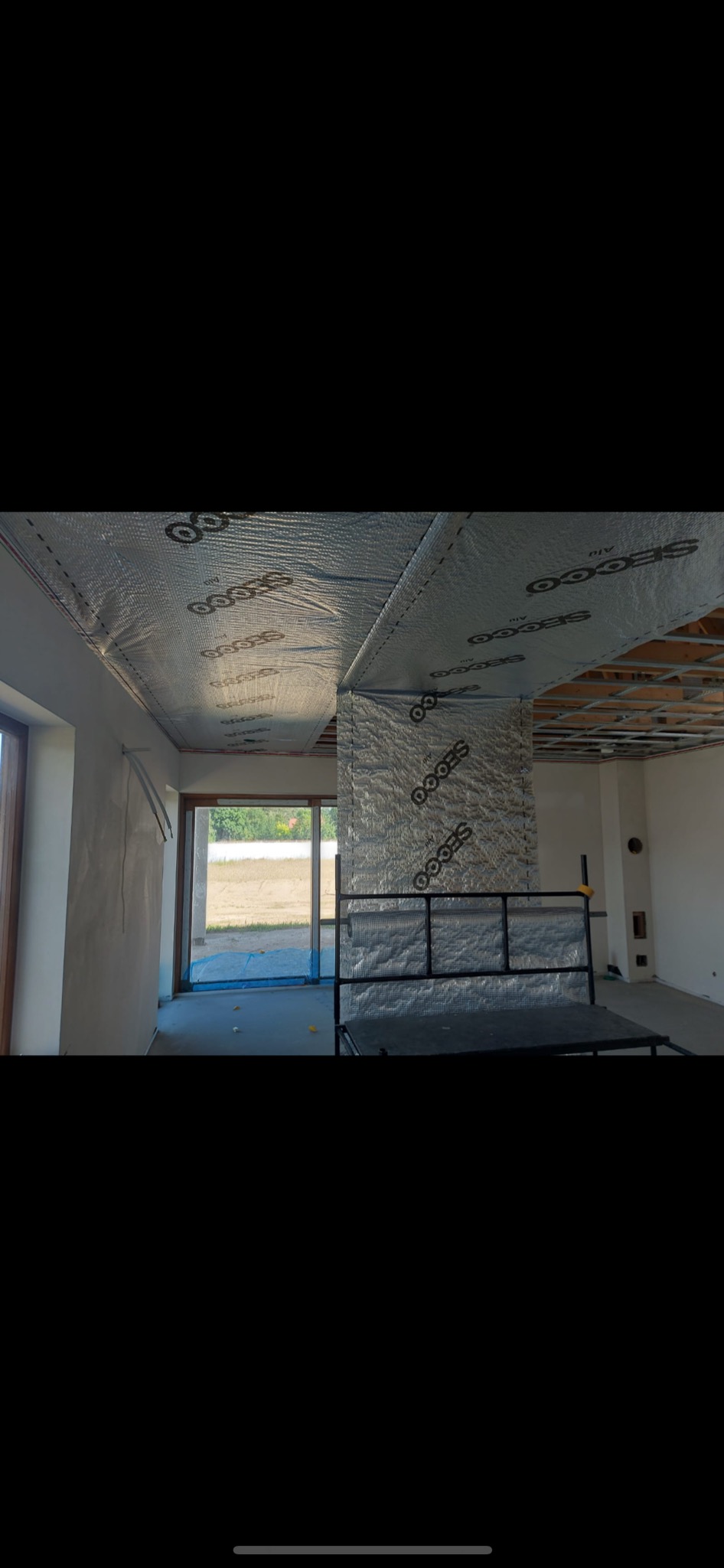 Wnętrze w trakcie budowy, widoczna izolacja termiczna z folii aluminiowej z nadrukiem na suficie i ścianie, surowy stan wykończenia, metalowa konstrukcja sufitu podwieszanego.