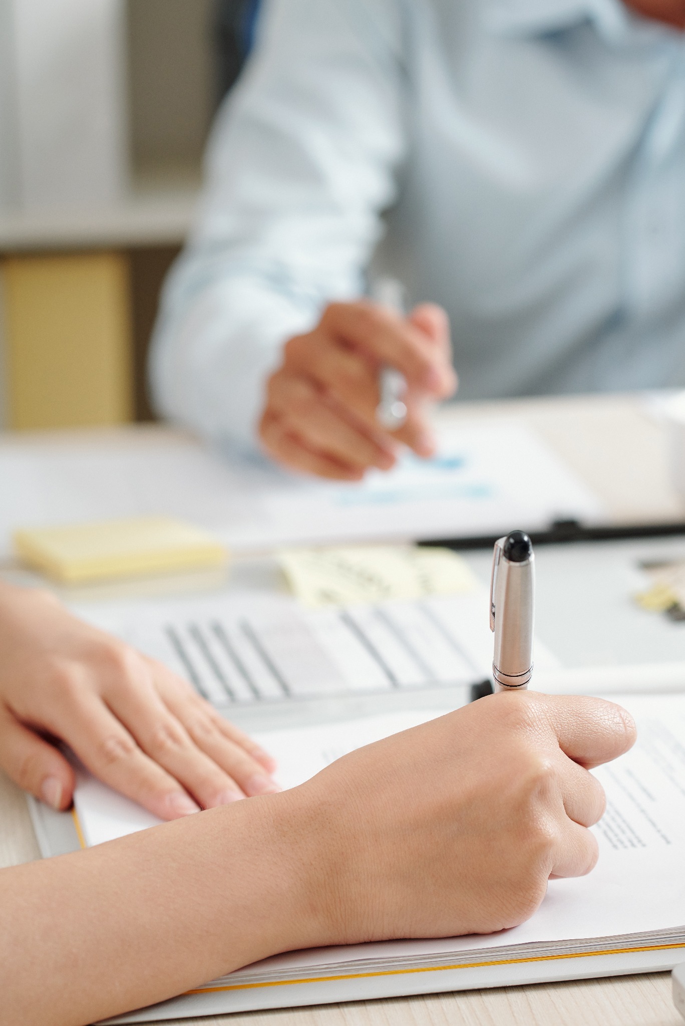 Osoba wypełnia dokument srebrnym długopisem, druga osoba w tle trzyma długopis nad papierami z wykresami.