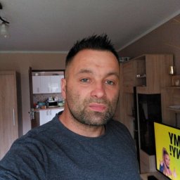 Tolux - Mężczyzna w szarej koszulce na tle wnętrza mieszkania z widoczną kuchnią i meblami.