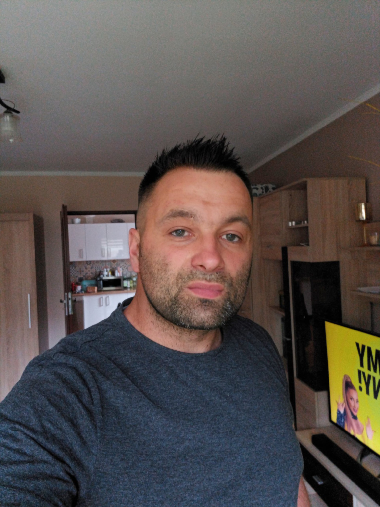 Mężczyzna w szarej koszulce na tle wnętrza mieszkania z widoczną kuchnią i meblami.