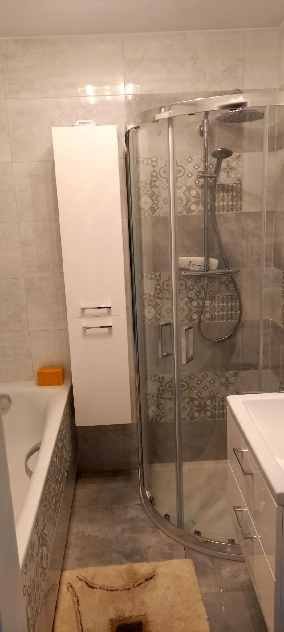 Wykończona łazienka z kabiną prysznicową narożną, białą szafką wiszącą z dwoma frontami, wanną z obudową dekoracyjną oraz beżowym dywanikiem na szarej podłodze.