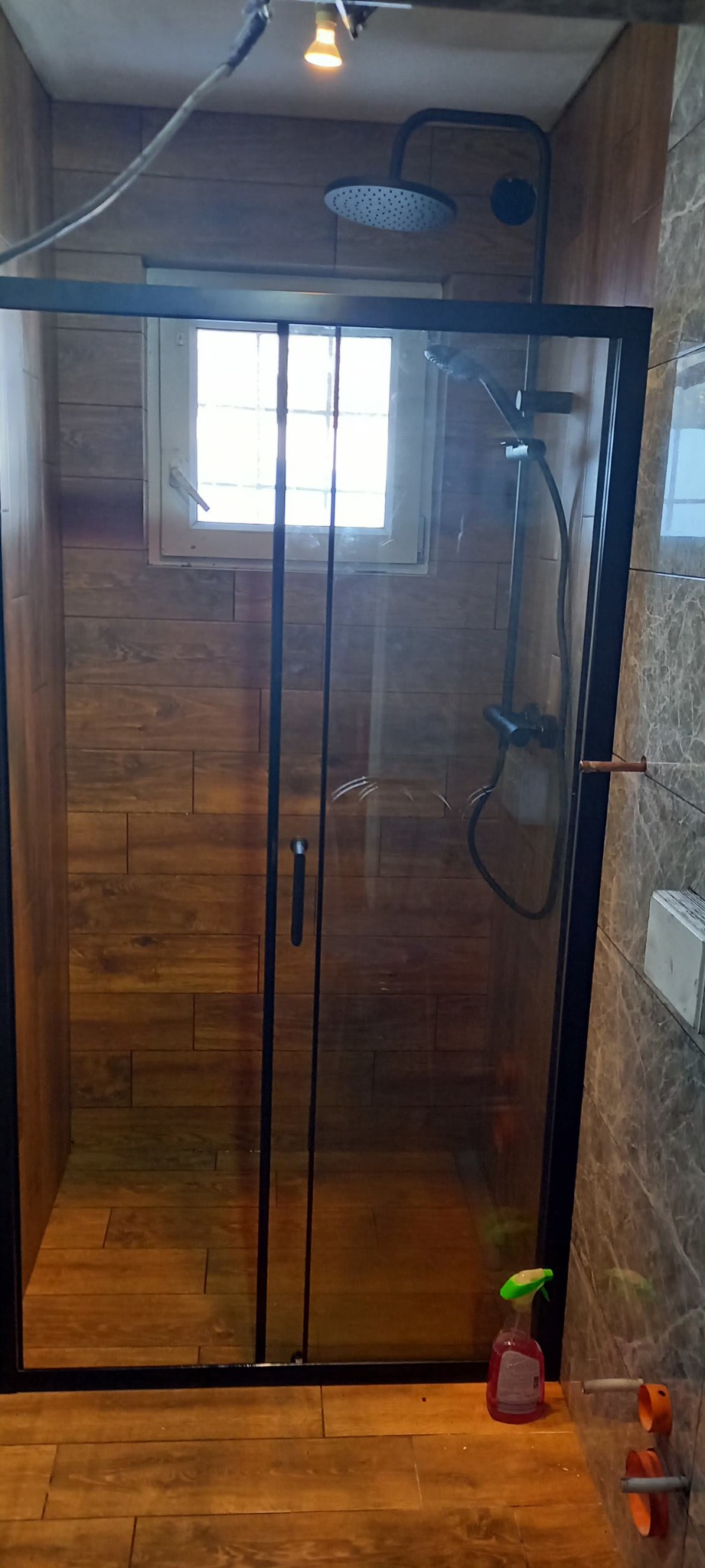 Kabina prysznicowa z czarną ramą i przesuwnymi drzwiami, drewnopodobne płytki na ścianach i podłodze, okno w tle, butelka środka czyszczącego w rogu.