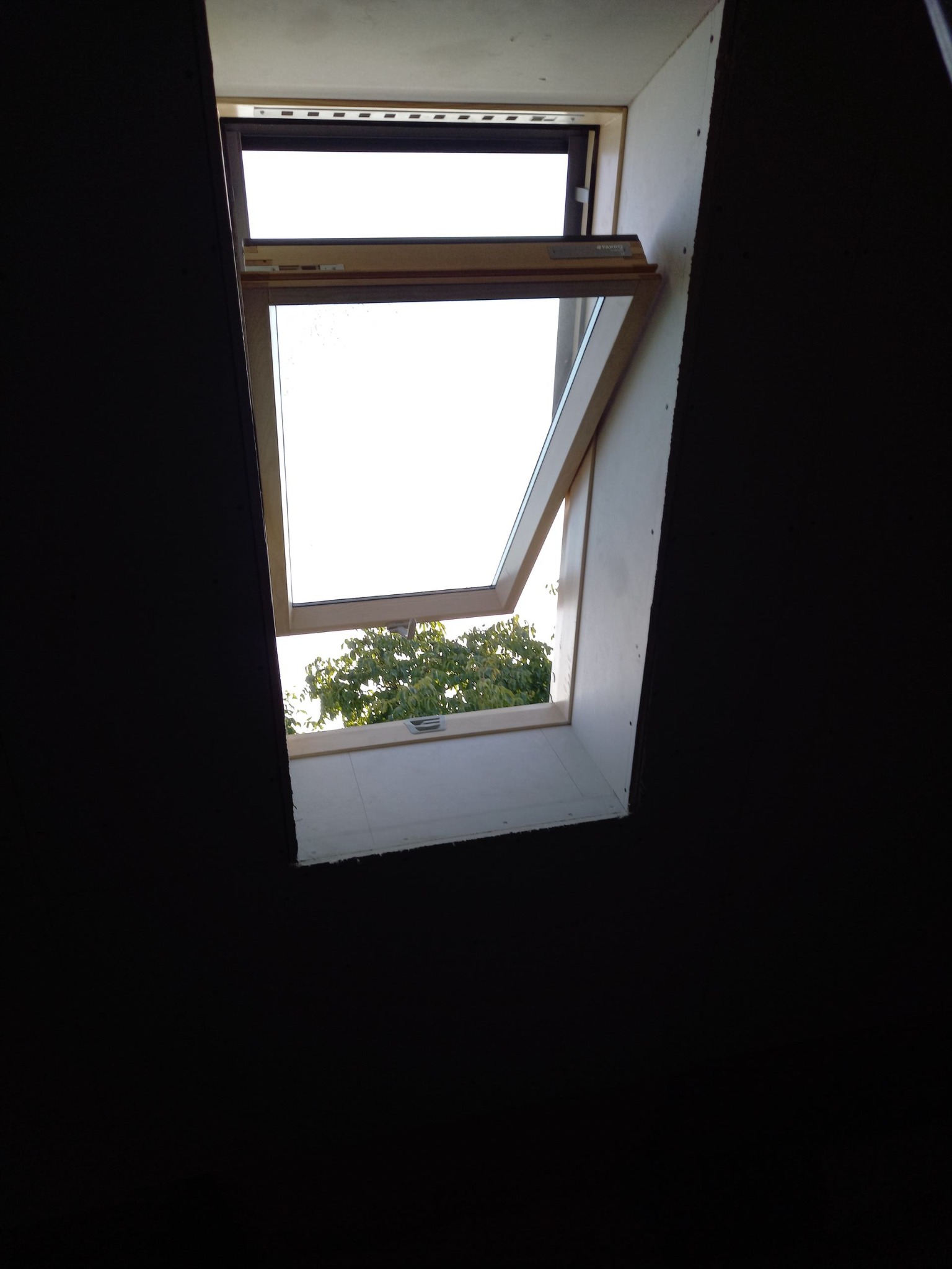 Otwarty okno dachowe z widokiem na zieleń drzew, osadzone w surowym wykończeniu ściany, z widocznymi śladami montażu.