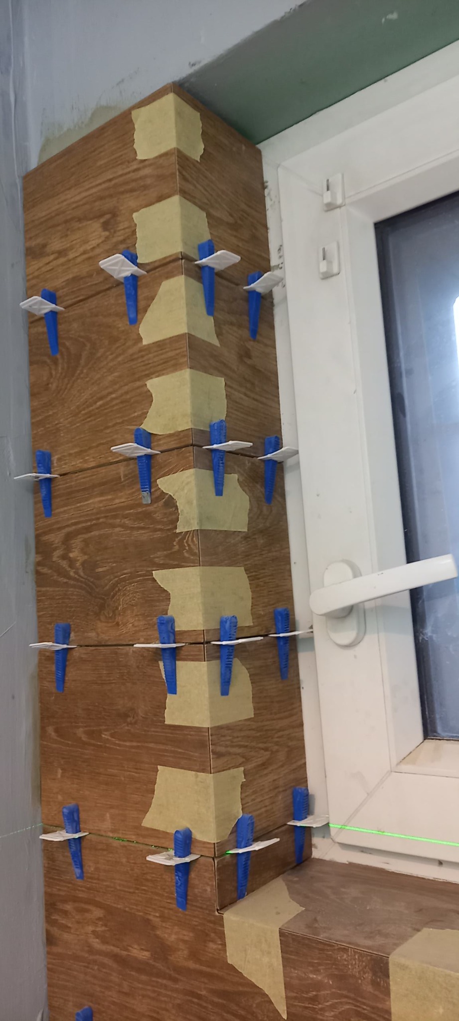 Układanie brązowych płytek imitujących drewno na narożniku ściany przy oknie, z użyciem niebieskich klinów i poziomicy laserowej.