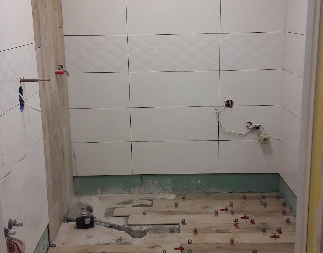 Łazienka w trakcie prac: białe płytki ścienne z delikatnym wzorem rombów, fragment ściany wyłożony płytkami imitującymi drewno, przygotowane podłoże z rozłożonymi płytkami podłogowymi i czerwonymi...