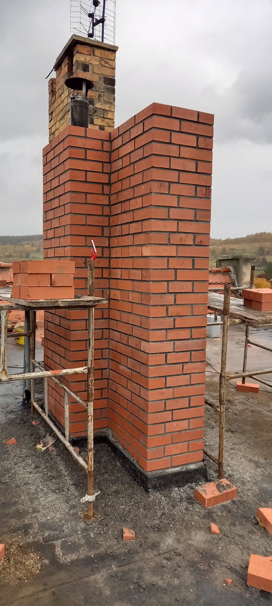 Budowa nowego komina z czerwonej cegły na dachu, widoczne rusztowanie i zapas cegieł, tło stanowią drzewa i pochmurne niebo.