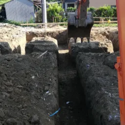 Pomarańczowa koparka w trakcie wykopu wąskich rowów pod fundamenty, widoczne ślady opon na ziemi i luźna ziemia na brzegach wykopu.