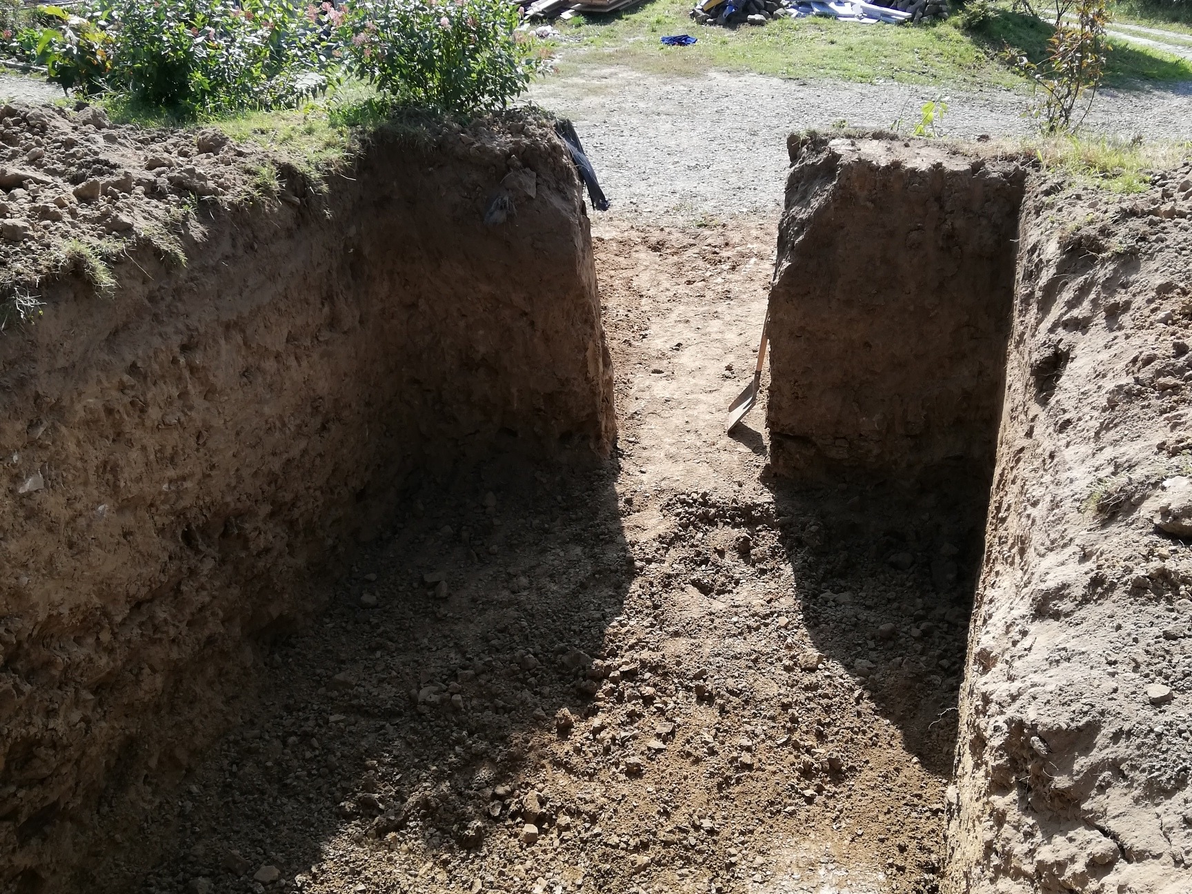 Głęboki wykop w ziemi z równymi ścianami, na dnie widoczne kamienie i grudki ziemi, w tle zieleń i fragment drogi, łopata oparta o jedną ze ścian.