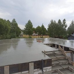 Zakład Ogólnobudowlany Jakub Szymkowiak - Świeżo wylany, mokry beton na fundamencie domu jednorodzinnego z widocznym szalunkiem schodów i wystającymi prętami zbrojeniowymi, tło stanowią drzewa i zabudowania.