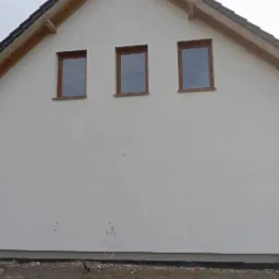 Boczny widok na nowo otynkowany dom z trzema oknami w drewnianych ramach, widoczne wystające kable i fragment gruntu.