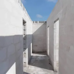 Wnętrze surowego, jasnego budynku w trakcie budowy z szarych bloczków, widoczne otwory drzwiowe i fragment nieba z chmurami.