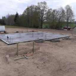 Zalany betonowy fundament z wystającymi prętami zbrojeniowymi na placu budowy, widoczne drewniane stemple i deski szalunkowe, w tle drzewa i budynek.